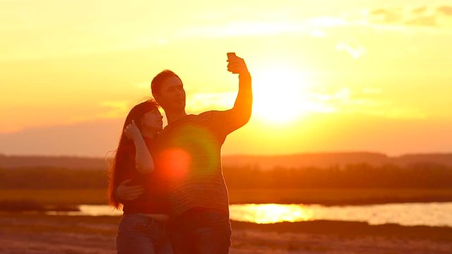 happy girl and guy doing selfie at sunset against a beautiful sky, they are happily in love, the girl's hair fluttering in the wind