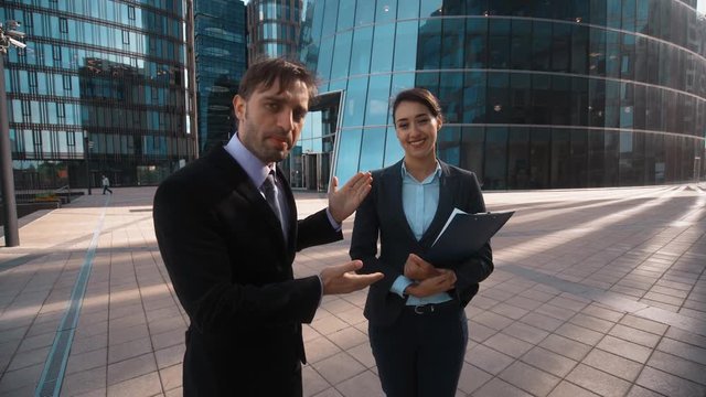 4K. UltraHD. POV. Young attractive Businessman in formal suit recommed his partner sexy pretty brownhair lady businesswoman with textpad to You. Modern glass building bg. Teal and orange sunset.