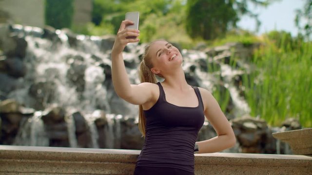 Young woman taking selfie in park. Selfie woman. Woman selfie. Girl taking selfie photo near waterfall. Blonde girl taking selfie on phone. Selfie girl taking photo outdoor. Sporty girl making selfie