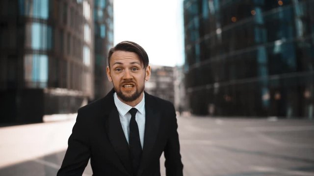 4k. UltraHD. POV. Portrait of angry Businessman swear and hate You. Formal suit and tie with beard and mustache. . Waving his arms, want to fight. Modern building glass district background. Teal