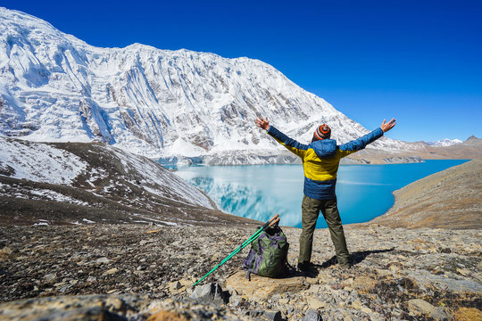 Travel concept: Happy hiker with rised hands, tourist sticks and backpack on Tilicho lake. Its 4900m above sea level. Snowly mountains and Tilicho lake on background.