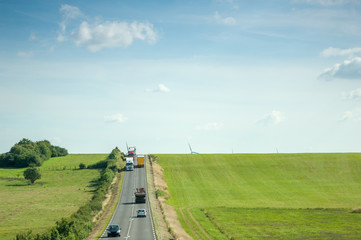 Aerial view of highway with two lane wih cars, trucks adn motorcycles driving between green fields and wind mills behind the hills