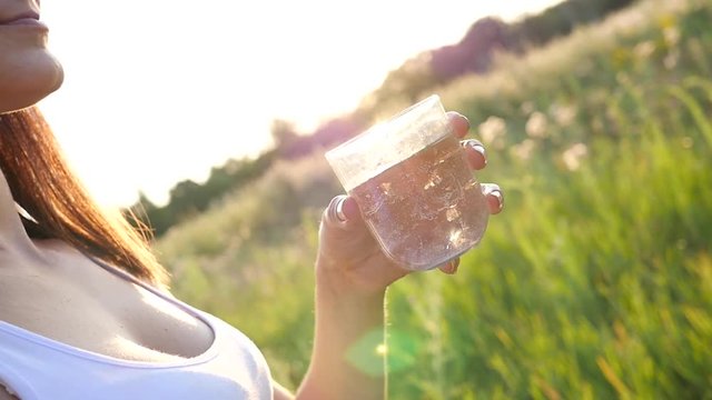 Young woman drinking clean pure water from glass on nature sunset light haze