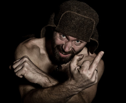 Dark portrait of scary evil sinister bearded man with smirk, showing middle finger. strange Russian man with a naked torso and a woolen hat.