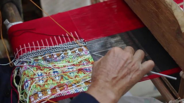 Old woman weaving red black and multi color cotton flag pattern on loom