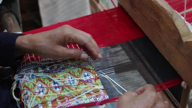 Old woman weaving red black and multi color cotton flag pattern on loom