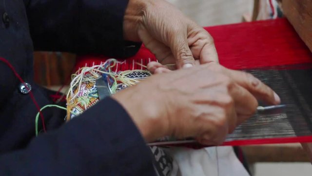 Old woman weaving red black and multi color cotton flag pattern on loom