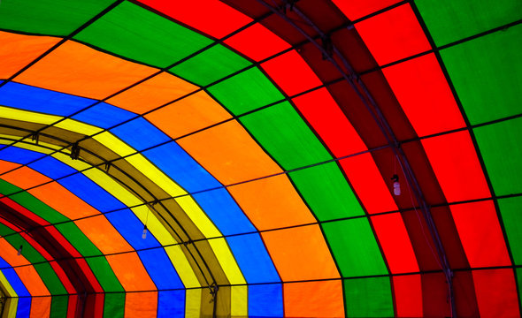 A photo taken inside a colorful tent. The tent consists of green, red, orange, blue and yellow colors and form repetitive patters of color strips. There are lamps hanging down the tent props.