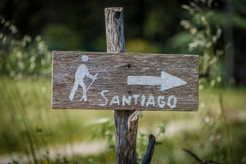 Schild aus Holz mit weisem Pfeil, Symbol eines Pilgers und Aufschrift Santiago am Jakobsweg in Spanien