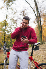 Obraz premium Autumn outdoors. Young smiled handsome guy with earphones standing with bicycle in a park, looking at cell phone and talking with someone.