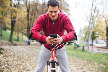Obraz premium Autumn outdoors. Young smiled handsome guy with earphones standing with bicycle in park, looking at cell phone and talking with someone.