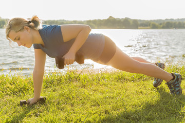 Young woman performing dumbbell row exercise lifting 12 pound weight in green grass outdoor park by...