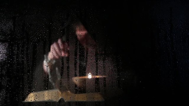 necromancer casts spells from thick ancient book by candlelight, behind transparent glass covered by water drops on a dark background