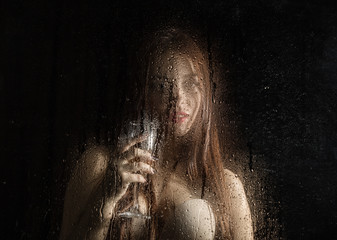 Smooth portrait of sexy model, posing behind transparent glass covered by water drops. young woman holding a glass of champagne