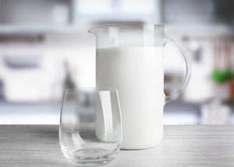 Empty glass with jug of milk on white wooden table against blurred kitchen background. Dairy concept.