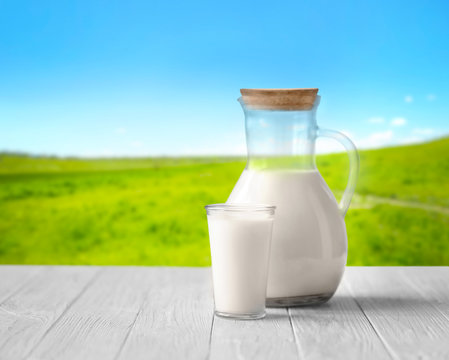Glass and jug with milk on white wooden table against blurred nature background. Dairy concept.