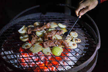 Making barbeque at night. Human hand holding fork arranging sausages, potatoes and onions on grill. Braai, outdoors activity in South Africa.