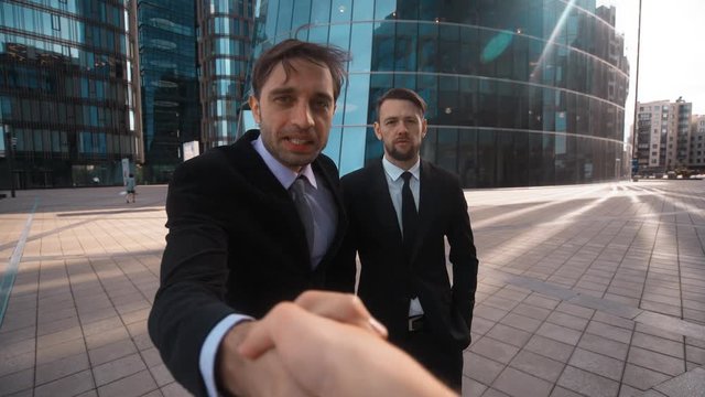 Young successful Businessman in formal suit recommed his confident collegue partner beard man in costume and tie to You. Modern glass business-centre building background. Teal and orange sunrise