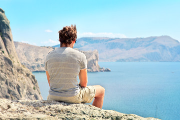 Naklejka na ściany i meble Young Man relaxing on rocky cliff sitting and looking on Sea and mountains Summer time Freedom concept
