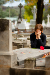 Distraught woman crying at graveside. She is covering her face with her hands. She has placed a single red flower on the stone grave.
