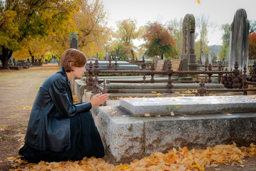 Young Woman Dressed in Black Praying at Grave in Cemetery in Fall