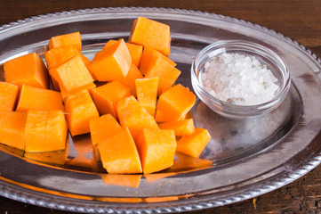 Slices of pumpkin cut into a metal tray. Salt in a bowl. On a wooden background.