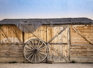Obraz premium wheel of an old chariot against the gate of a warehouse made of wood and a thatched roof