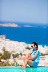 Fototapeta na wymiar Slim woman applying sunscreen on her legs, sitting on the edge of pool background old town Mykonos in Europe