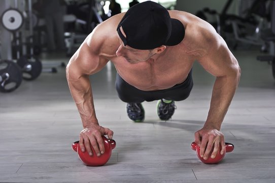 Handsome powerful athletic man performing push ups with kettle bell. Strong bodybuilder with  perfect shoulders, biceps and triceps.