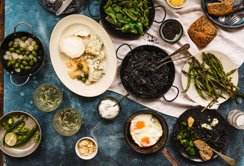 Rich vegetarian dinner with black pasta, grilled green vegetables, cheese and fried eggs over blue table. 