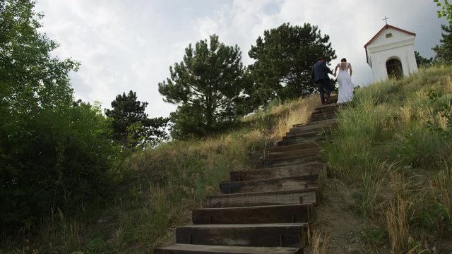 A newly married couple climbs a staircase to a shrine with a cross above.  They pause for a kiss near the top.  4k. In slow motion.