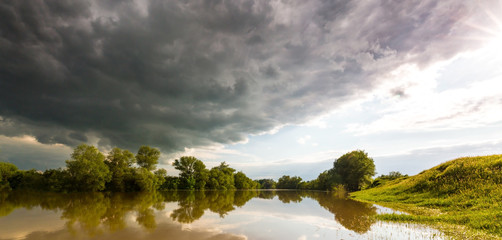 Ominous stormy sky over natural flooded river, with bright sun emerging from under the cumulus cloud cover