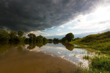 Ominous stormy sky over natural flooded river, with bright sun emerging from under the cumulus...