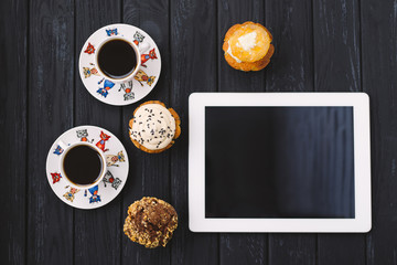 three homemade muffins, two cups of coffee and a tablet on black wooden background
