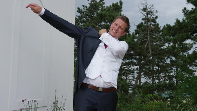 A groom puts on his blazer. A forest and white shrine are behind him. 4k. In slow motion.