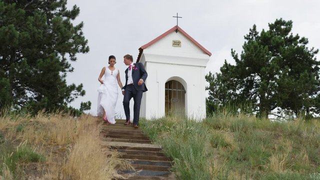 A newly married couple descends a staircase with a shrine and cross behind them.  They pause for a kiss.  4k. In slow motion.