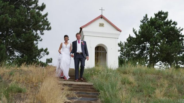 A newly married couple descends a staircase with a shrine and cross behind them. 4k. In slow motion.