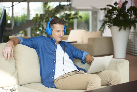 Relaxed weekend. Shot of a middle aged man with his laptop sitting on sofa at home and listening music.