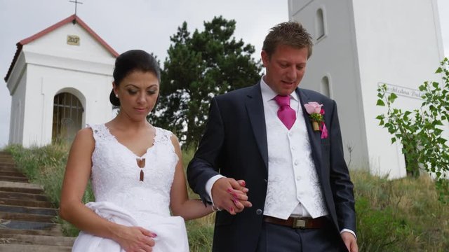 A newly married couple descends a staircase while holding hands with a shrine and cross behind them. 4k. In slow motion.
