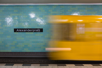 Gelbe U-Bahn in Bewegung. Berlin Alexanderplatz-Schild an der Wand der U-Bahn-Station sichtbar.