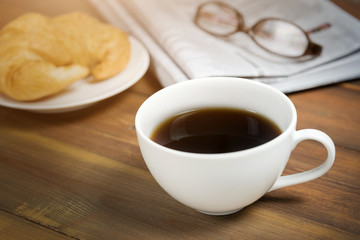Coffee cup with newspaper and glasses in background on wooden table.