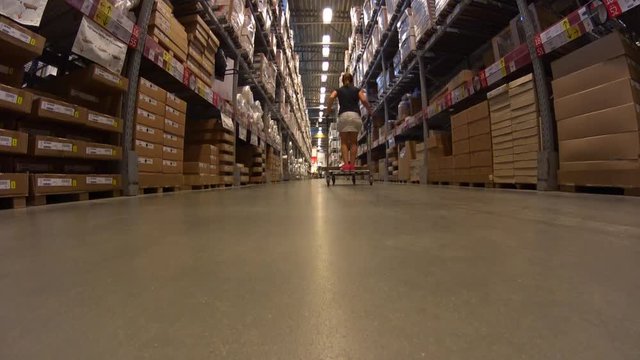 Funny Young Girl in mini skirt rides shopping cart, go the distance in furniture warehouse store between rows of shelves with cardboard boxes
