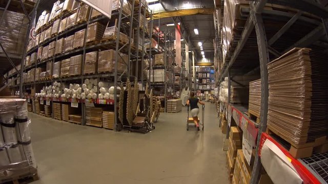 Young woman rides shopping trolley in furniture warehouse and faced with shelf with cardboard boxes