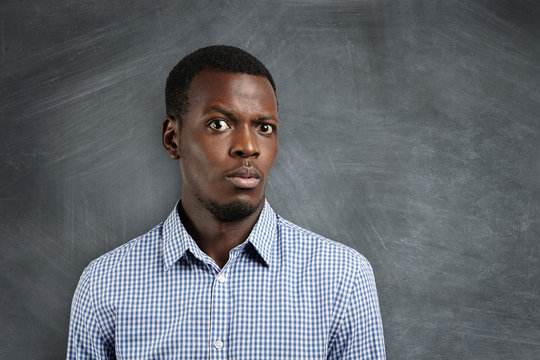 Human face expressions and emotions. Close up shot of doubtful African man dressed casually looking confused and puzzled, raising his brow in surprise and indignation, standing at blank blackboard