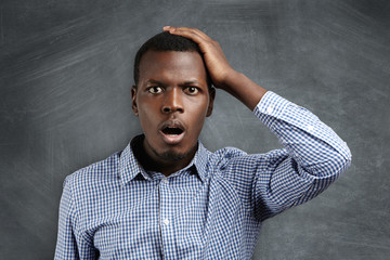 Portrait of forgetful young dark-skinned customer or employee wearing checkered shirt looking with...