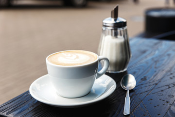 A white cup of cappuccino, sugar bowl and a tea spoon on the dark wooden table of a city cafe terrace