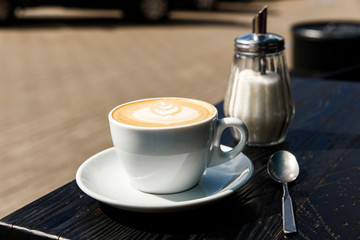 A white cup of cappuccino, sugar bowl and a tea spoon on the dark wooden table of a city cafe...
