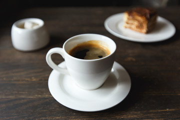 A white cup of black coffee, a piece of cake on the white plate, sugar bowl on the wooden table in a cafe. Selective focus, small depth of fieild.
