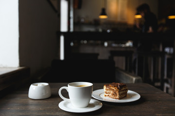 A white cup of black coffee, a piece of cake on the white plate, sugar bowl on the wooden table in a cafe. Selective focus, small depth of fieild.
