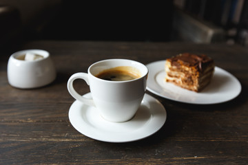 A white cup of black coffee, a piece of cake on the white plate, sugar bowl on the wooden table in a cafe. Selective focus, small depth of fieild.